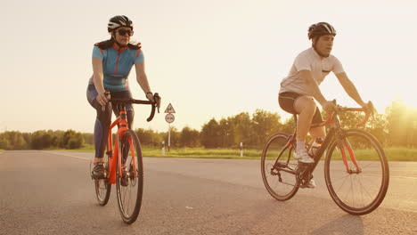 Zwei-Radfahrer,-Ein-Mann-Und-Eine-Frau,-Fahren-Mit-Helmen-Und-Sportbekleidung-Bei-Sonnenuntergang-In-Zeitlupe-Auf-Rennrädern-Auf-Der-Autobahn